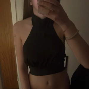Svart halterneck croppad topp - Säljer en svart croppad halterneck topp i glansigt material och fina mönster. Toppen knyts i nacken och bak på ryggen vilket ger en öppen rygg. Perfekt för dig som vill ha en stilren och trendig look. Toppen har gått sönder lite i nacken men den funkar bra ändå och går lätt att fixa där av priset🥰pris går att diskutera