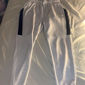 Grå mjukisbyxor från McKenzie med svarta detaljer på sidan  - Snygga grå mjukisbyxor från McKenzie med svart längs sidorna och Byxorna har elastisk midja, perfekt för en avslappnad stil. Märkeslogga på vänster lår. Hör av er vid frågor 