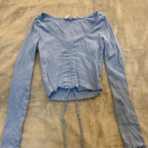 Ljusblå croppad topp med knyt - Säljer en ljusblå långärmad topp med v-ringning och knyt framtill. Toppen är croppad och har en ribbad struktur samt vågig kant nertill. Perfekt för dig som gillar trendiga och bekväma plagg.