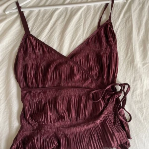 Vinröd omlottopp med smala axelband - Säljer en vinröd omlottopp med smala axelband och knyt i sidan. Toppen har plisserat tyg och en glansig finish som ger en lyxig känsla. Perfekt för dig som vill ha något snyggt och trendigt!