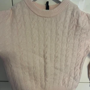 Ljusrosa kabelstickad tröja - Säljer en ljusrosa kabelstickad tröja med rund halsringning och korta ärmar. Tröjan har ett klassiskt flätmönster framtill och ribbade muddar vid hals och midja. Perfekt för lager-på-lager och en söt vardagslook.
