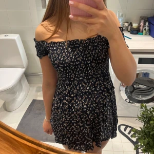 Blommig offshoulder klänning från Brandy Melville - Supersöt kort offshoulder klänning från Brandy Melville i mörkblått med små vita och beige blommor. Smockad överdel och volangkant nertill. Perfekt för sommarens alla tillfällen!