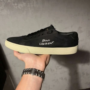 Saint Laurent Svarta Sneakers - Snygga svarta sneakers från Saint Laurent med vit sula och diskret logga på sidan. Skorna har snörning och är perfekta för en stilren look. Tillverkade i ett slitstarkt material som ger en lyxig känsla. 