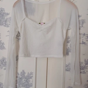 Vit croppad topp med lång ärm och blomdetalj - Säljer en söt vit croppad topp med lång ärm och en liten rosa blomdetalj framtill. Toppen har en smockad textur och är perfekt för en stilren look. Passar bra till både jeans och kjol!