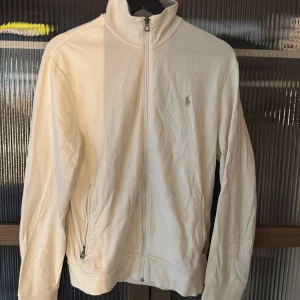 Vit/beige hoodie från Polo Ralph Lauren - Säljer en stilren vit/beige tröja från Polo Ralph Lauren. Den har en dragkedja framtill och långa ärmar. Den är perfekt för en avslappnad look och har en broderad logga på bröstet. Köpt för 2800kr