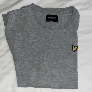 Grå t-shirt från Lyle & Scott - Säljer en stilren grå t-shirt från Lyle & Scott med korta ärmar och ett broderat emblem på bröstet. Perfekt för en avslappnad look. Skön och enkel att matcha med det mesta. Hör av dig vid intresse!