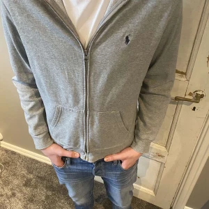 Grå hoodie från Ralph Lauren - Säljer en stilren grå hoodie från Ralph Lauren med dragkedja och det klassiska logotypen på bröstet. Perfekt för en avslappnad look. Tröjan har långa ärmar och en bekväm passform. Priset är ej hugget i sten, kontakta oss vid funderingar!🤝