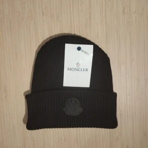 Svart ribbad mössa från Moncler - Snygg svart ribbad mössa från Moncler med broderad logga framtill. Perfekt för att hålla värmen under kyliga dagar. Mössan har en klassisk design med uppvikt kant.