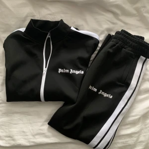 Svart tröja och byxor från Palm Angels - Snygg svart träningsjacka från Palm Angels med vita ränder längs ärmarna och dragkedja framtill. Jackan har en hög krage och broderad logga på bröstet. Perfekt för en sportig look!
