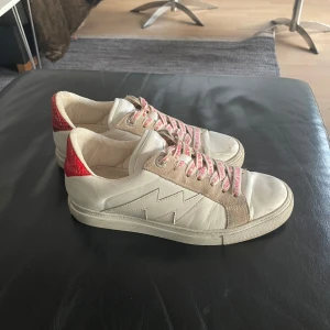 Vita sneakers från Zadig & Voltaire - Snygga vita sneakers från Zadig & Voltaire med röda detaljer och snörning. Skorna har en unik design med röd ormskinnsmönstrad häl och rosa snören med logotyp. Perfekta för en trendig look. Använda men bra skick, lite repor vid framsidan❤️