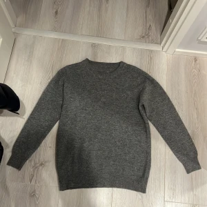 Grå stickad tröja - Säljer en stilren grå stickad tröja med rund hals och långa ärmar. Perfekt för kyligare dagar och enkel att matcha med olika outfits. Tröjan har ribbade muddar vid ärmslut och nederkant.