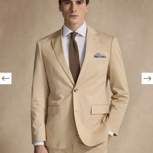 Beige blazer från These Glory Days -48 - Elegant beige blazer från These Glory Days med en klassisk kavaj och matchande byxor. Kostymen har en stilren design med två knappar framtill och en bröstficka med en dekorativ näsduk. Perfekt för formella tillfällen.