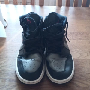 Svarta och grå Nike Air Jordans - Säljer ett par klassiska Nike Air Jordans i svart och grått med röd logga. Skorna har snörning och en hög design som ger extra stöd. Perfekta för dig som älskar streetwear och vill ha en tidlös stil.