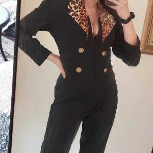 Svart jumpsuit med leopardmönstrad krage - Snygg svart jumpsuit med leopardmönstrad krage och guldknappar framtill. Perfekt för en djärv och stilren look. 