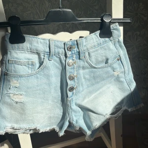 Ljusa jeansshorts från Zara - Säljer ett par ljusa jeansshorts från Zara med slitna detaljer och fransar vid bensluten. Shortsen har knappar framtill och klassiska femficksdesign. Perfekta för en avslappnad stil!