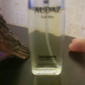 Audaz Eau de Parfum - Säljer en elegant flaska Audaz Eau de Parfum från Insieme. Flaskan är genomskinlig med en svart kork och innehåller en gyllene doftvätska. Perfekt för den som söker en sofistikerad och unik doftupplevelse.