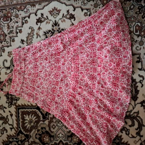 Röd och vit blommig kjol - Charmig långkjol med rött blommönster på vit bakgrund och små guldprickar. Perfekt för en somrig look med sitt lätta och luftiga material.