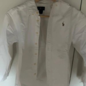 Vit skjorta från Ralph Lauren - Klassisk vit skjorta från Ralph Lauren med broderad logga på bröstet. Skjortan har långa ärmar och knappar framtill. Perfekt för en stilren look.