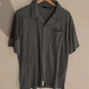 Grå kortärmad skjorta från 157 - Säljer en stilren grå kortärmad skjorta från 157. Skjortan har en klassisk krage och knappar framtill. Den är tillverkad i ett mjukt material och har en bröstficka för extra stil. Perfekt för en avslappnad look. Passar även bra nu när sommaren börjar närma sig! Pris kan förhandlas