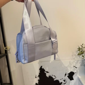 Ljuslila axelväska från Gymshark - Säljer en ljuslila axelväska från Gymshark. Väskan har ett stilrent och kompakt format med dragkedja upptill och breda axelband. Perfekt för att bära med sig det nödvändigaste. Den har även en praktisk innerficka med dragkedja för extra säkerhet. Säljer då jag köpt en annan väska