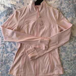 Ljusrosa jacka från Lululemon - Säljer en ljusrosa jacka från Lululemon med dragkedja framtill och hög krage. Jackan har långa ärmar och är tillverkad i ett mjukt, stretchigt material. Perfekt för träning eller en avslappnad dag. Den har också praktiska fickor med dragkedjor. Lappen är avklippt men i övrigt är den i nyskick 