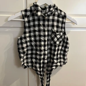Svartvit rutig ärmlös knytblus från H&M - Säljer en cool svartvit rutig ärmlös blus från H&M. Blusen har knappar framtill och en knytning i nederkant för en avslappnad look. Perfekt för varma dagar eller som ett lager under en jacka.