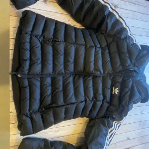 Säljer en svart dunjacka från Adidas med klassiska vita ränder på ärmarna. Jackan har en dragkedja framtill och är långärmad. Perfekt för kyliga dagar med sin pufferdesign som håller värmen. Adidas-logga på bröstet ger en sportig touch.