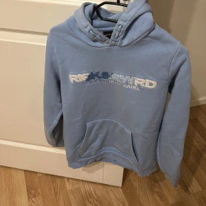 Ljusblå risk and reward tröja - Säljer en ljusblå hoodie med broderad text på framsidan. Tröjan har en stor ficka framtill och en bekväm huva. Perfekt för en avslappnad stil.