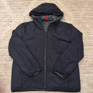 Svart jacka från Polo Ralph Lauren - Säljer en svart jacka från Polo Ralph Lauren med en broderad röd logga på bröstet. Jackan har en dragkedja framtill och en huva. Perfekt för kyligare dagar.