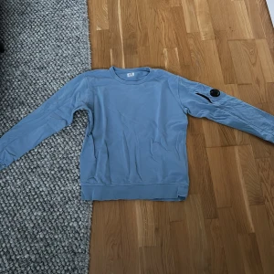 Blå sweatshirt från C.P. Company - Säljer en snygg blå sweatshirt från C.P. Company. Tröjan har långa ärmar och en rund halsringning. På ärmen finns en cool detalj med en ficka och en rund logotyp. Perfekt för en avslappnad stil. Pris går att diskuteras.