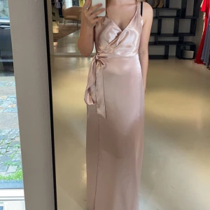 Ljusrosa balklänning  - Elegant  långklänning i satin med omlott-design och knytning i midjan. Klänningen har en v-ringad urringning och smala axelband, perfekt för balen