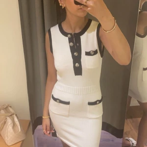 Vit ärmlös klänning med knappar - Snygg vit ärmlös klänning med svarta detaljer och stora knappar framtill. Den har en tight passform och ribbad midja för en smickrande siluett. Perfekt för en stilren look.