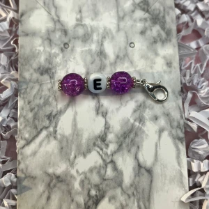 Nyckelring med lila pärlor och bokstav - Charmig nyckelring med två glittriga lila pärlor och en vit pärla med bokstaven 'E'. Utrustad med en praktisk karbinhake för enkel fastsättning.