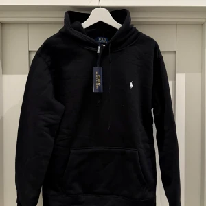 Svart hoodie från Polo Ralph Lauren - Säljer en stilren svart hoodie från Polo Ralph Lauren med det klassiska logotypbroderiet på bröstet. Tröjan har en bekväm passform med långa ärmar och en praktisk känguruficka framtill. Perfekt för en avslappnad look. Storlek M