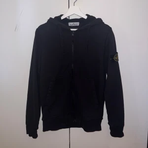 Svart hoodie från Stone Island - Säljer en stilren svart hoodie från Stone Island med dragkedja och huva. Den har en ikonisk logga på ärmen och är perfekt för en casual look. Passar bra till både jeans och joggers.