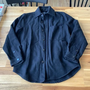Svart overshirt från A Day's March - Säljer en stilren svart overshirt från A Day's March i storlek S. Jackan är tillverkad av 80% ull och 20% polyamid, vilket ger en skön och varm känsla. Den har knappar framtill och en bröstficka för extra stil. Perfekt för lager-på-lager under kyligare dagar. Inga defekter jättebra skick!
