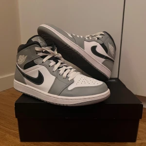 Nike Air Jordan 1 Mid Light Smoke Grey - Snygga Nike Air Jordans i en stilren kombination av grått och vitt i mycket bra skick och har aldrig använts mer än fem gånger. Skorna har en klassisk design med svart swoosh och snörning. Perfekta för dig som vill ha en ikonisk sneaker 
