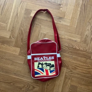 Röd axelväska med Beatles-tryck - Snygg röd axelväska med ett coolt Beatles-tryck på framsidan. Väskan har vita detaljer och en justerbar axelrem. Perfekt för musikälskare som vill ha en unik accessoar. Det har ett mindre fack innuti! 