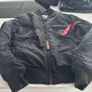 Svart bomberjacka från Alpha Industries - Säljer en klassisk svart bomberjacka från Alpha Industries med orange foder. Jackan har dragkedja och ribbade muddar vid ärmslut och nederkant. Den har en ikonisk 'Remove Before Flight'-tag på ärmen. Perfekt för en cool och avslappnad stil.