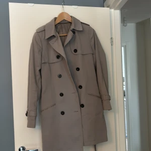 Grå  trenchcoat från Tiger of Sweden - Snygg ljusgrå trenchcoat från Tiger of Sweden i modellen Dorine. Jackan har en klassisk design med dubbelknäppning och bälte i midjan. Tillverkad i bomull med viskosfoder för en bekväm känsla. Perfekt för vår och höst. Passar om man brukar ha typ storlek 38. Extra knappar finns kvar. Kemtvätt. Katt finns i hemmet. Sista bilden visar färg i dagsljus. 