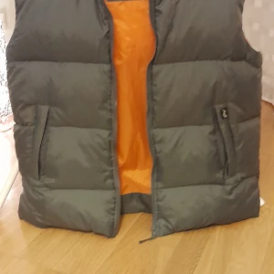Grå dunväst från Stuff Clothing - Säljer en grå dunväst från Stuff Clothing med en cool orange insida. Västen har en dragkedja framtill och är perfekt för kyligare dagar. Den är stilren och enkel att matcha med olika outfits.