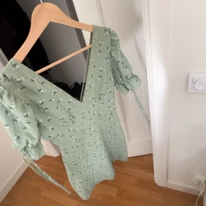 Grön blommig klänning - Söt grön klänning med blommigt mönster och v-ringning. Den har korta puffärmar med knytband och en smickrande passform. Perfekt för vår och sommar!