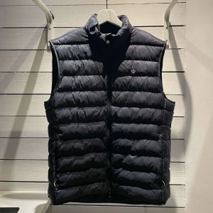 Svart dunväst från Morris - Säljer en stilren svart dunväst från Morris med dragkedja framtill. Västen har en quiltad design och är perfekt för kyligare dagar. Den är i storlek medium och passar bra för en klassisk look.