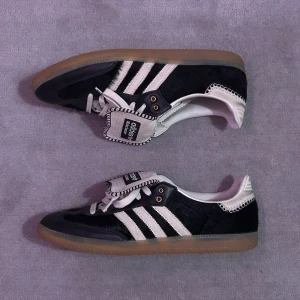 Adidas Wales Bonner Pony Samba sneakers - Storlek US 11.5. Nypris är över 10 000 SEK. Snygga Adidas Wales Bonner sneakers i svart och vitt med klassiska tre ränder på sidan. Skorna har en unik design med en bred flik över snörningen och en gummisula i brunt. Perfekta för dig som vill ha en stilren och sportig look.