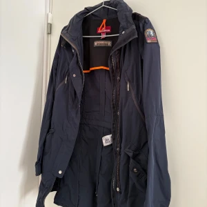 Mörkblå jacka från Parajumpers vindjacka - Säljer en mörkblå jacka från Parajumpers med dragkedja och knappar. Jackan har en patch på ärmen och är perfekt för blåsiga dagar. Den har flera praktiska fickor och en justerbar huva.
