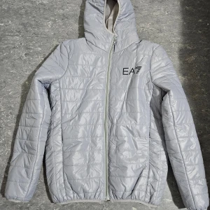 Grå quiltad jacka från EA7 - Säljer en stilren grå quiltad jacka från EA7. Jackan har en dragkedja framtill och en praktisk huva. Perfekt för kyligare dagar.