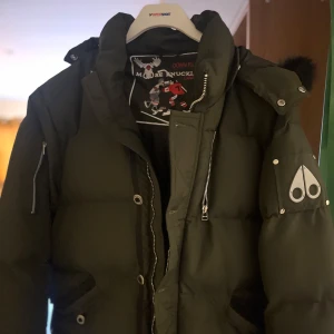 Dunjacka från Moose Knuckles - Säljer en dunjacka från Moose Knuckles med en stilren design. Jackan har en dragkedja framtill och flera knappar. Den är långärmad och har en hög krage för extra värme. Perfekt för kyliga dagar. Använd endast 2 gånger. Storlek L