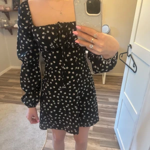 Svart blommig klänning - Säljer en söt svart klänning med vita blommor. Klänningen har långa ärmar och en knytning framtill. Perfekt för en stilren look.