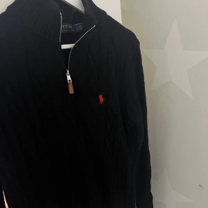 Svart stickad tröja från Ralph Lauren - Snygg svart stickad tröja från Ralph Lauren med dragkedja och röd logga på bröstet. Perfekt för kyligare dagar med sin ribbade krage och stilrena design. Passar både till vardags och mer uppklädda tillfällen.