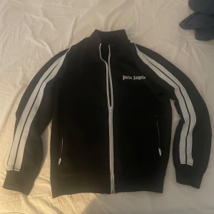 Svart track jacket från Palm Angels - Säljer en svart track jacket från Palm Angels med vita ränder längs ärmarna och dragkedja framtill. Jackan har en broderad logotyp på bröstet och är tillverkad i Italien. Perfekt för en sportig look!